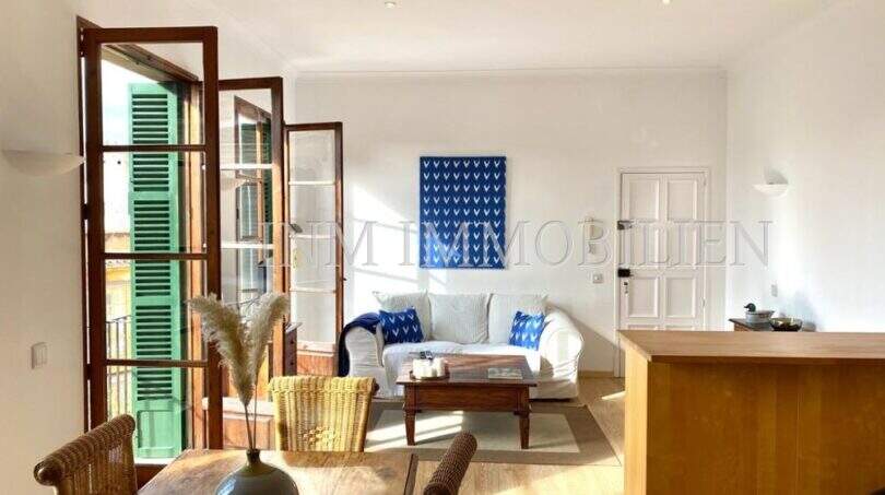 Sala de estar luminosa con sofá blanco, suelo de madera y contraventanas verdes típicas en apartamento del casco antiguo de Palma