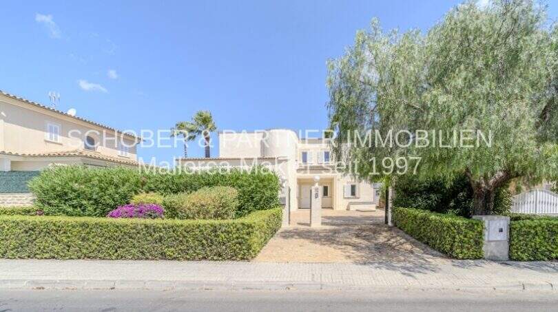 Außenansicht mediterrane Villa in Sa Coma mit gepflegtem Grüngarten, Einfahrt und Palmen unter blauem Himmel.
