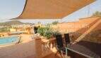 Covered terrace with sun sail, dining table, chairs and view of pool and hills in Son Servera