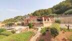 Exterior view Finca Son Servera: Mediterranean garden with pool, natural stone house and wooded hill backdrop