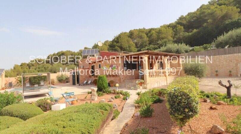 Exterior view Finca Son Servera: Mediterranean garden with pool, natural stone house and wooded hill backdrop