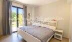 Helles Schlafzimmer mit weißem Doppelbett, Nachttisch und Terrassenzugang zum Garten in Eck-Wohnung Sa Coma