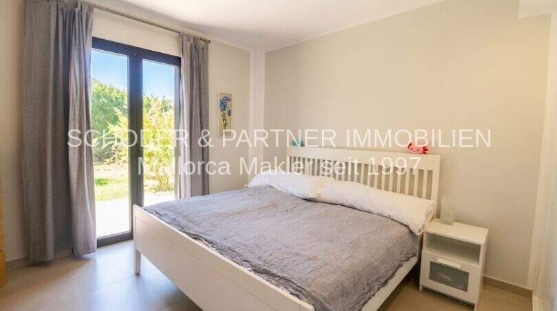 Helles Schlafzimmer mit weißem Doppelbett, Nachttisch und Terrassenzugang zum Garten in Eck-Wohnung Sa Coma