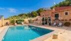 Exterior view of Finca Son Servera with turquoise-blue pool, natural stone house, sun terrace and loungers under blue sky.