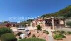 Exterior view of Finca Son Servera: Mediterranean garden, pool, natural stone house with terrace and solar system in front of wooded hills.