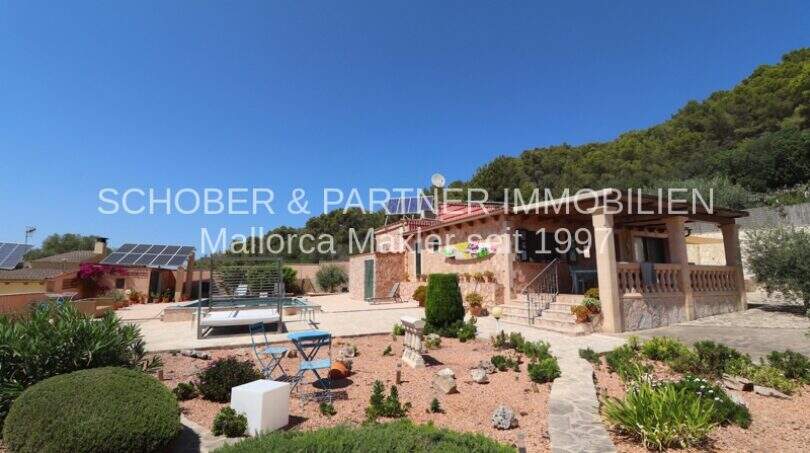 Exterior view of Finca Son Servera: Mediterranean garden, pool, natural stone house with terrace and solar system in front of wooded hills.