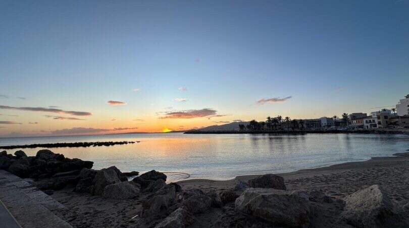 Sonnenuntergang über dem Meer am Strand von Portitxol, Palma de Mallorca, mit Palmen und Stadtsilhouette
