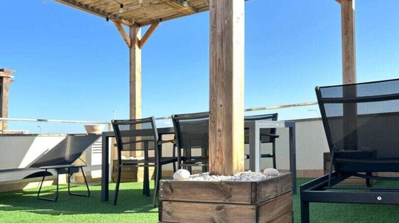 Dachterrasse mit Holzpergola, Lichterketten, Kunstrasen, Loungemöbeln und Esstisch unter blauem Himmel.