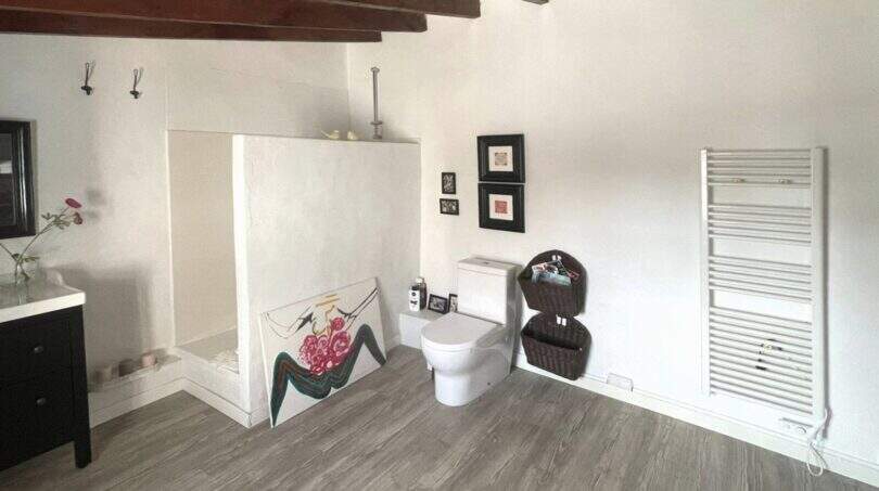 Baño espacioso con techo de vigas de madera, paredes blancas, WC, toallero calefactor y obra de arte en la casa de pueblo Sencelles