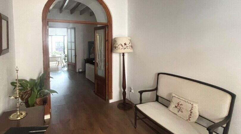Pasillo elegante con arco redondo, techo de vigas de madera, banco tapizado color crema y lámpara de pie en la casa de pueblo Sencelles
