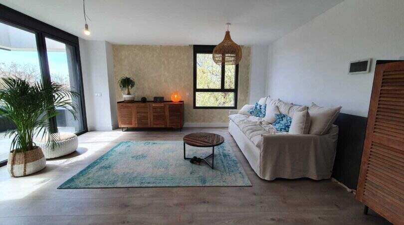 Helles Wohnzimmer des Penthouses in Nou Llevant mit Holzboden, weißem Sofa, Rattanlampe und großer Fensterfront.