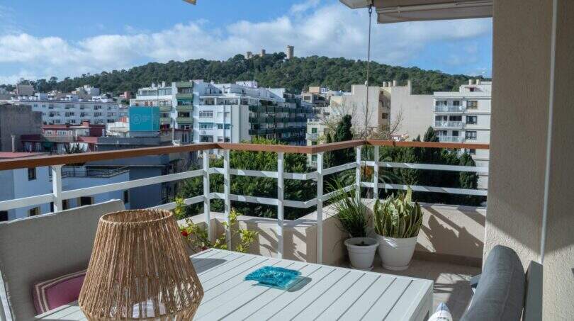 Sonnige Dachterrasse mit Stadtblick auf Palma, weißem Tisch, Rattanlampe und Bellver-Burg im Hintergrund