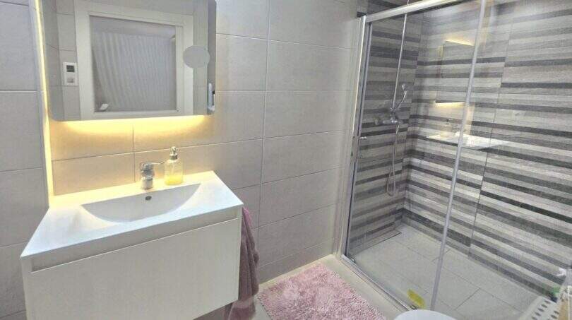 Modern bathroom with illuminated mirror, white sink and glass shower in Palmanova apartment