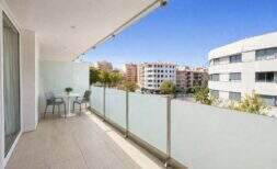 Sonniger Balkon mit Glasgeländer, Tisch und Stühlen, Blick auf Palmas Stadtbild und blauer Himmel