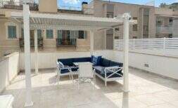 Soleada terraza en la azotea con pérgola blanca, mobiliario lounge azul y vistas a la ciudad en Palma de Mallorca