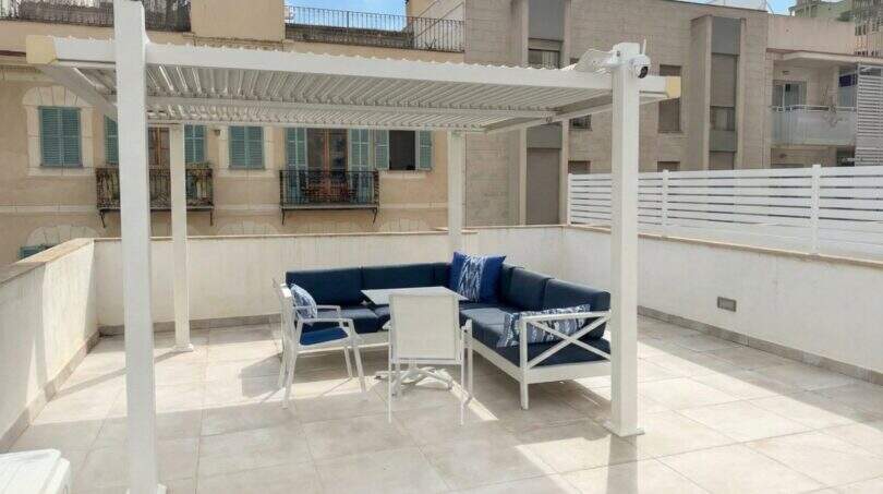 Soleada terraza en la azotea con pérgola blanca, mobiliario lounge azul y vistas a la ciudad en Palma de Mallorca