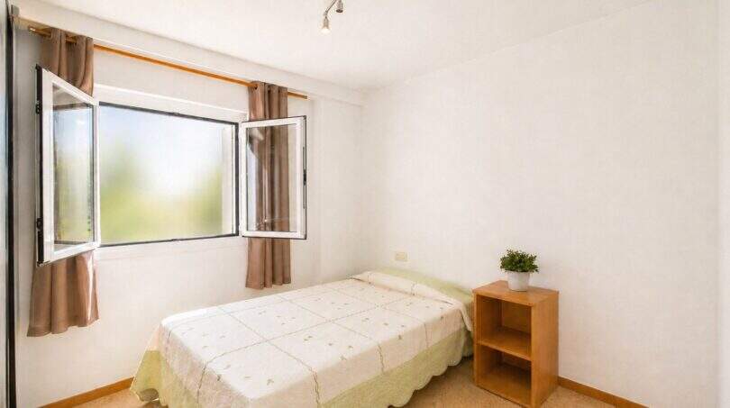 Helles Schlafzimmer der Wohnung in Portixol mit Einzelbett, Holzregal und geöffnetem Fenster mit Gardinén.