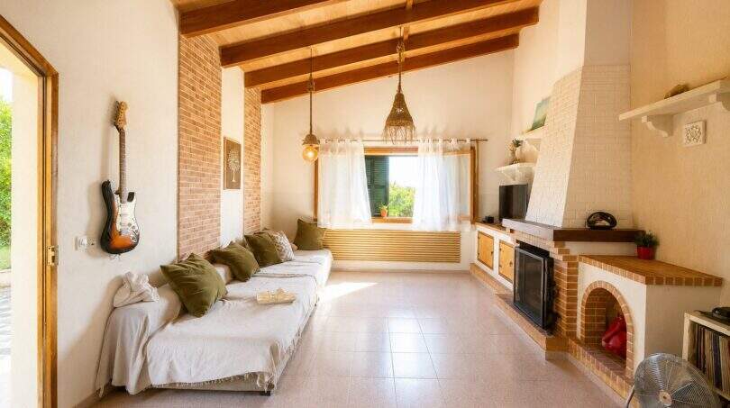 Sala de estar luminosa de una finca mallorquina con techo de vigas de madera, sofá blanco, chimenea y guitarra en la pared.