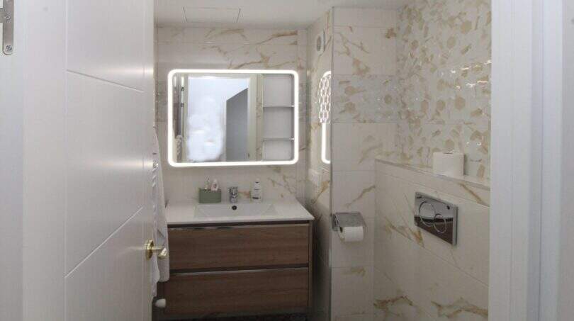 Modernes Badezimmer mit beleuchtetem Spiegel, Holzwaschtisch und Marmoroptik-Fliesen im Reihenhaus Palma.