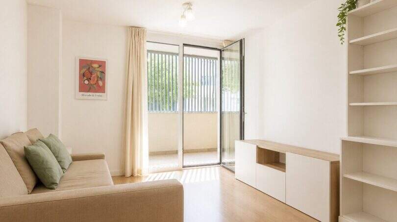 Helles Wohnzimmer der Palma-Wohnung mit Balkonzugang, Holzboden, Bücherregal und beigem Sofa mit grünen Kissen.