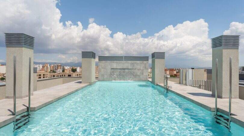 Infinity-Pool auf dem Dach mit Wasserfall und Stadtpanorama von Palma de Mallorca unter blauem Himmel.