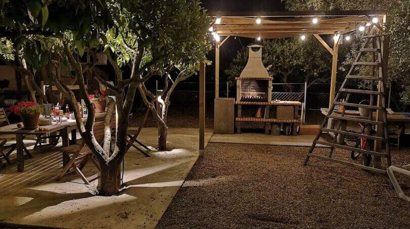 Abendliche Gartenanlage der Finca in Campanet mit beleuchtetem Pergola-Grillbereich und Sitzecke unter Bäumen.