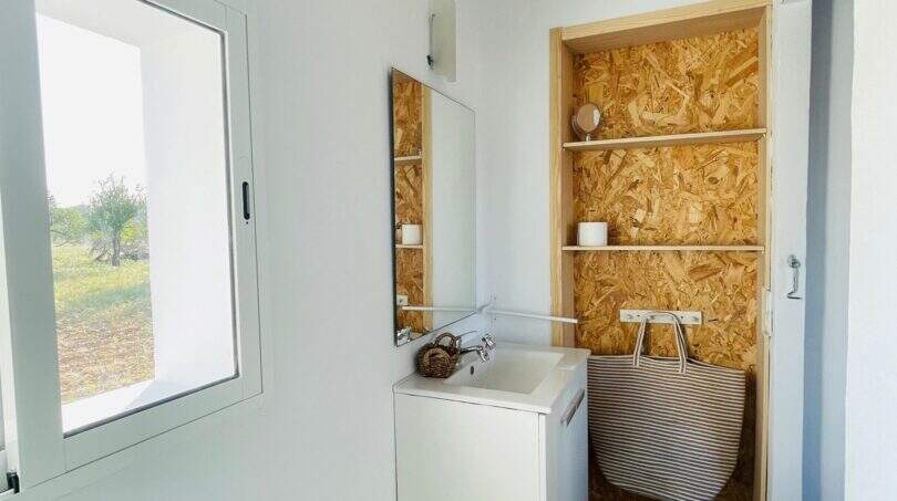 Rustikales Badezimmer der Finca in Campanet mit Holzbalken, OSB-Regalwand, Waschbecken und Fensterblick ins Grüne.
