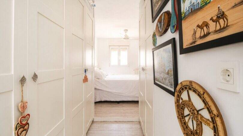 Heller Flur mit weißen Einbauschränken, Holzboden und Blick ins weiße Schlafzimmer, dekoriert mit Gemälden und Wanduhr.