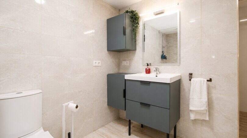 Modernes Badezimmer der Finca bei Palma mit grauem Waschtischunterschrank, Wandspiegel und hellen Marmorfliesen.