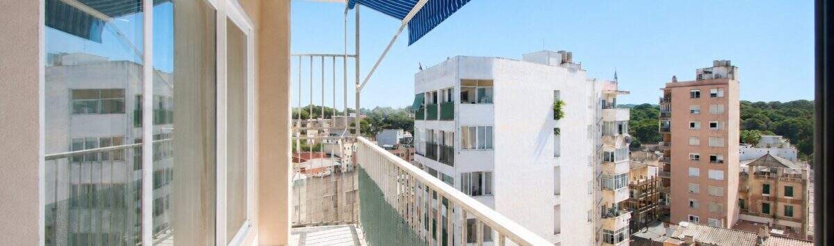 Sonniger Balkon mit blauem Sonnensegel und Stadtblick auf Palma de Mallorca, helle Glasfront