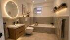Modernes Badezimmer mit Badewanne, Waschtisch, beleuchtetem Rundspiegel und natürlichen Holzelementen in S'Arenal
