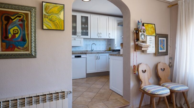 Wohnbereich mit Rundbogen zur weißen Küche, Terrakottaboden, Kunstwerken und Holzstühlen in Villa Marratxí