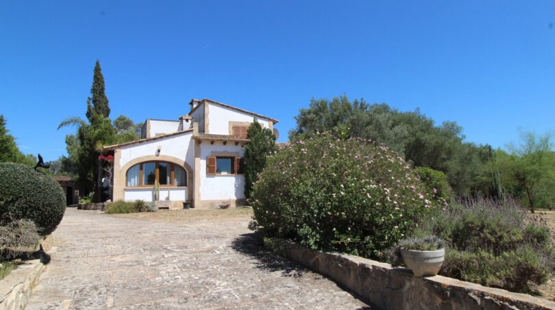 Außenansicht Villa Marratxí: weißes Landhaus mit Rundbogenfenster, Natursteineinfahrt und mediterranem Garten bei blauem Himmel.