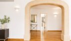 Zona de estar clara de la villa en Llucmajor con elegante arco de piedra arenisca, parquet de madera y vista a habitaciones adyacentes.