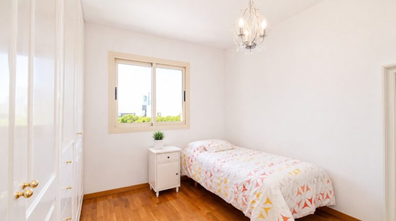Dormitorio luminoso de la villa en Llucmajor con cama individual, suelo de madera, lámpara de araña y ventana con vista al jardín.
