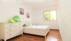 Dormitorio luminoso de la villa Llucmajor con cama blanca, cómoda, suelo de madera y ventana hacia el verde.