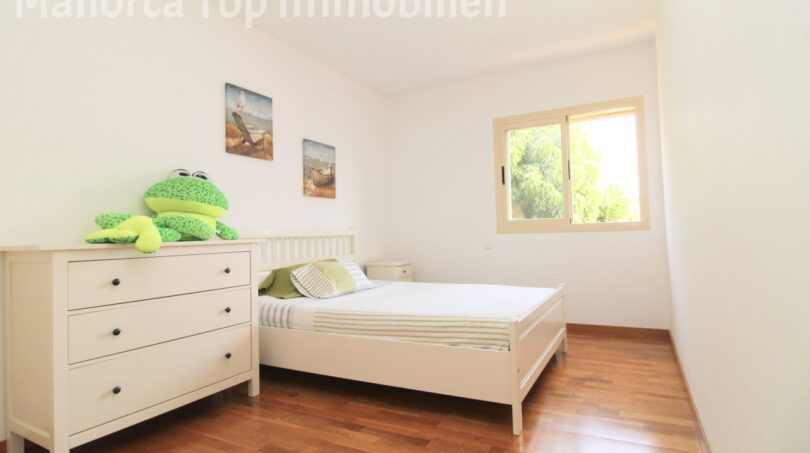 Dormitorio luminoso de la villa Llucmajor con cama blanca, cómoda, suelo de madera y ventana hacia el verde.