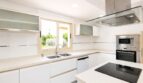 Moderna cocina blanca de la villa en Llucmajor con isla de cocción, electrodomésticos integrados y ventana hacia el jardín