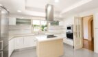 Moderna cocina blanca de la villa en Llucmajor con isla de cocción, campana extractora de acero inoxidable y electrodomésticos integrados.