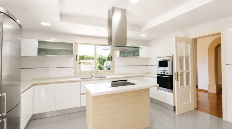 Moderna cocina blanca de la villa en Llucmajor con isla de cocción, campana extractora de acero inoxidable y electrodomésticos integrados.