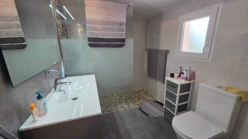 Modernes Badezimmer der Finca S'Illot mit Walk-in-Dusche, Waschtisch, WC und Fenster in Grautönen.