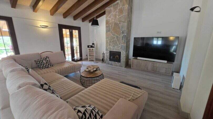 Helles Wohnzimmer der Finca mit Holzbalkendecke, Steinwand, Kamin und großem TV in S'Illot