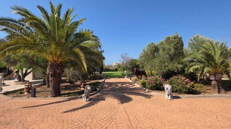 Gepflasterte Einfahrt mit Palmen, Löwenskulpturen und roter Stierfigur im mediterranen Garten der Finca S'Illot