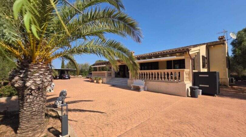 Finca S'Illot: Gepflasterter Innenhof mit Palmen, Steinbalustrade und mediteraner Fassade unter blauem Himmel.