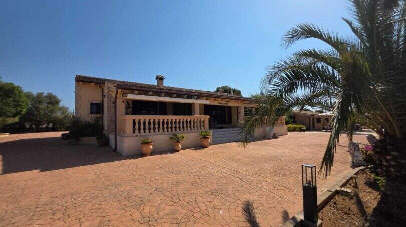Außenansicht der Finca in S'Illot mit gepflastertem Vorplatz, weißer Veranda und Palmen unter blauem Himmel.