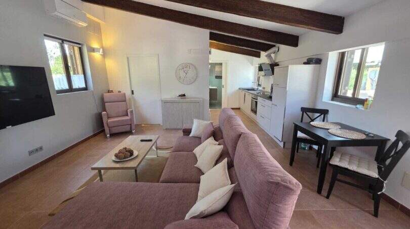 Helles Wohnzimmer der Finca S'Illot mit Holzbalkendecke, rosa Sofa, offener Küche und Klimaanlage