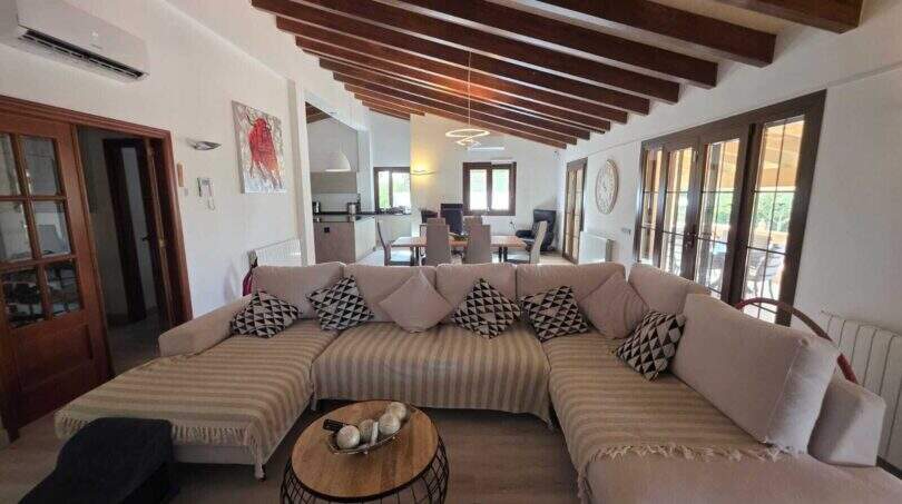 Helles Wohnzimmer der Finca S'Illot mit großem Ecksofa, Holzbalkendecke und offenem Zugang zur Küche