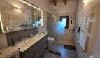 Modernes Badezimmer der Finca S'Illot mit Doppelwaschbecken, beleuchtetem Spiegel, Dusche und Holzbalkendecke.