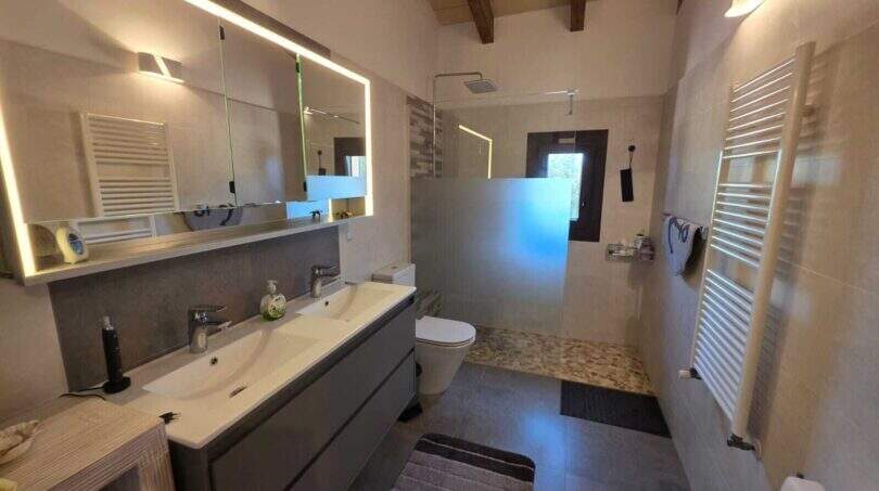 Modernes Badezimmer der Finca S'Illot mit Doppelwaschbecken, beleuchtetem Spiegel, Dusche und Holzbalkendecke.