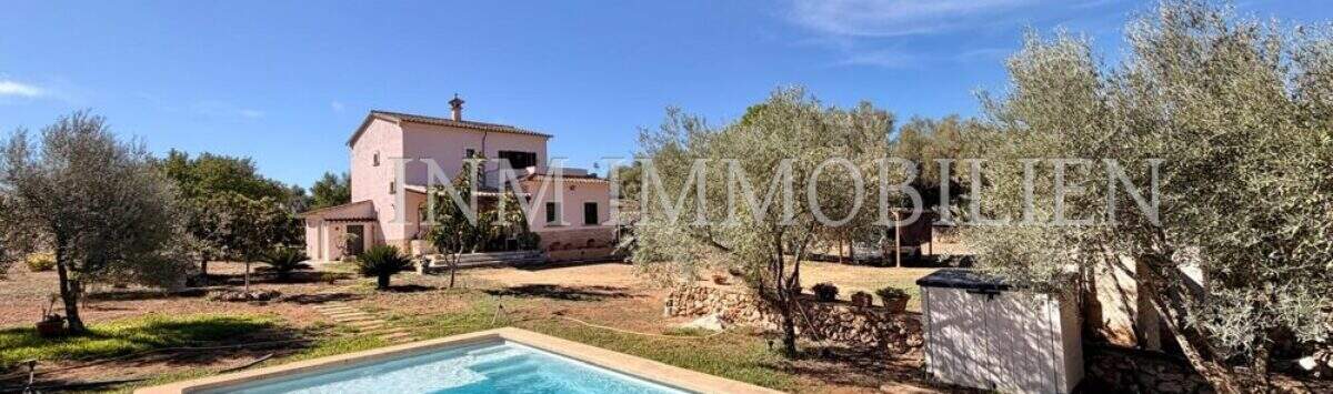 Außenansicht mediterrane Finca Algaida mit Pool, Olivenbäumen und rosa Haupthaus unter blauem Himmel.